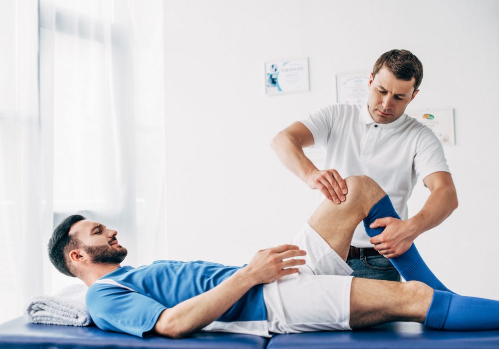Physiotherapist massaging leg of football player lying on massage table in hospital